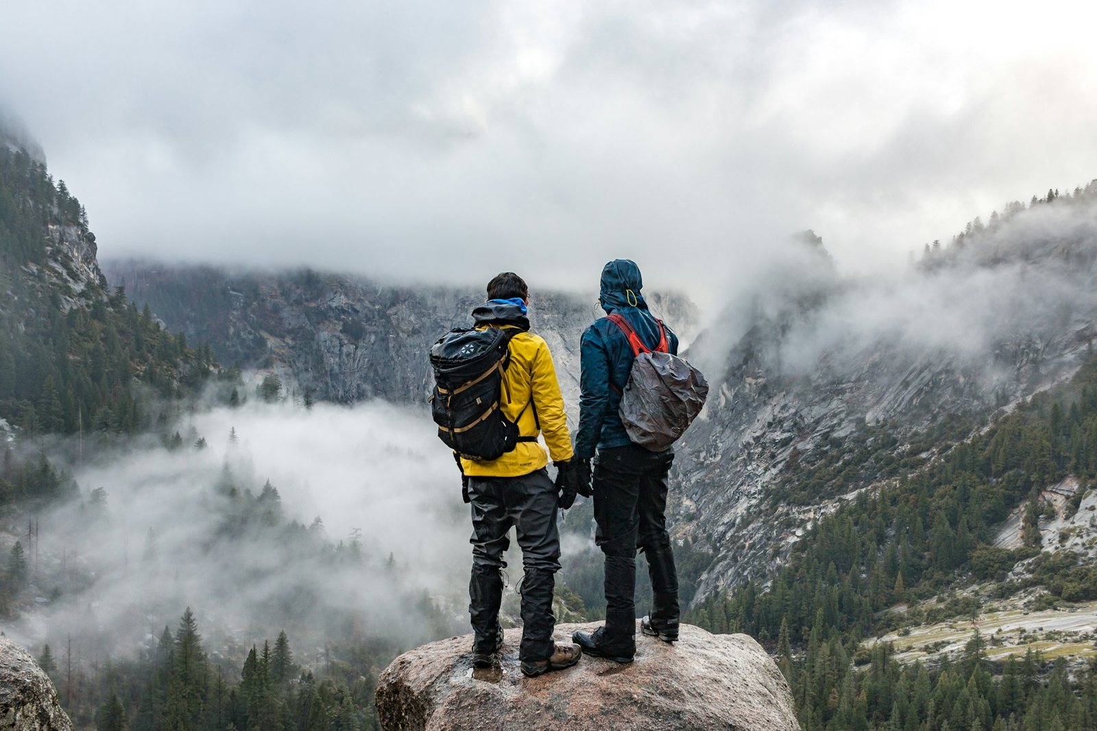 Dos senderistas con ropa impermeable y mochilas frente a un valle con niebla
