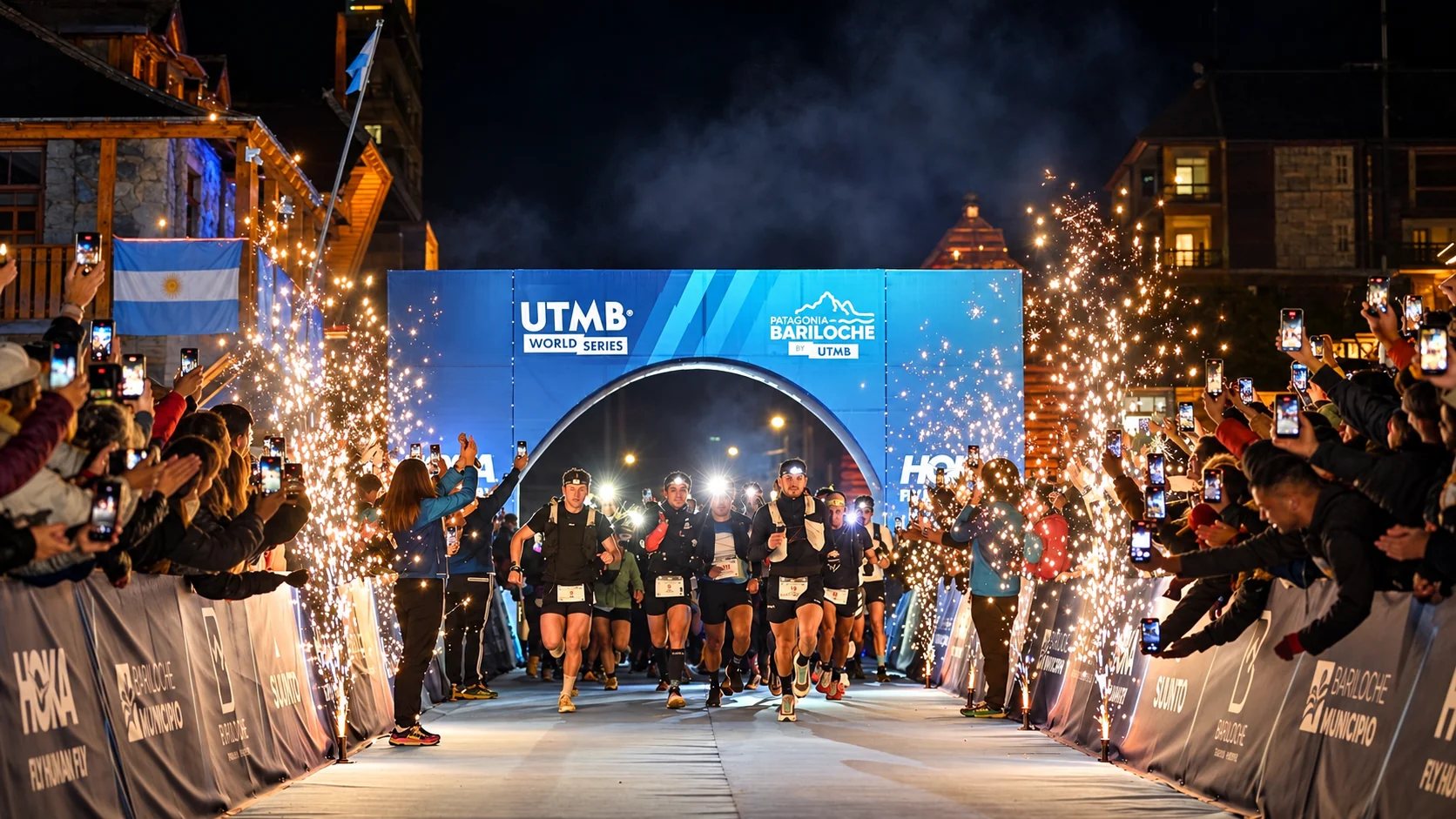Largada nocturna del UTMB Patagonia Bariloche: corredores con linternas bajo el arco de partida, entre el público y chispas de fuegos fríos